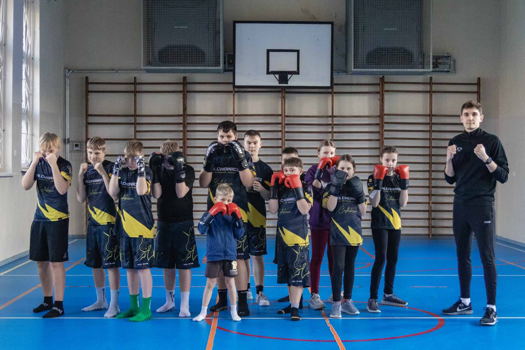 Grupa dzieci i młodzieży z trenerem stoi w gardzie na sali gimnastycznej